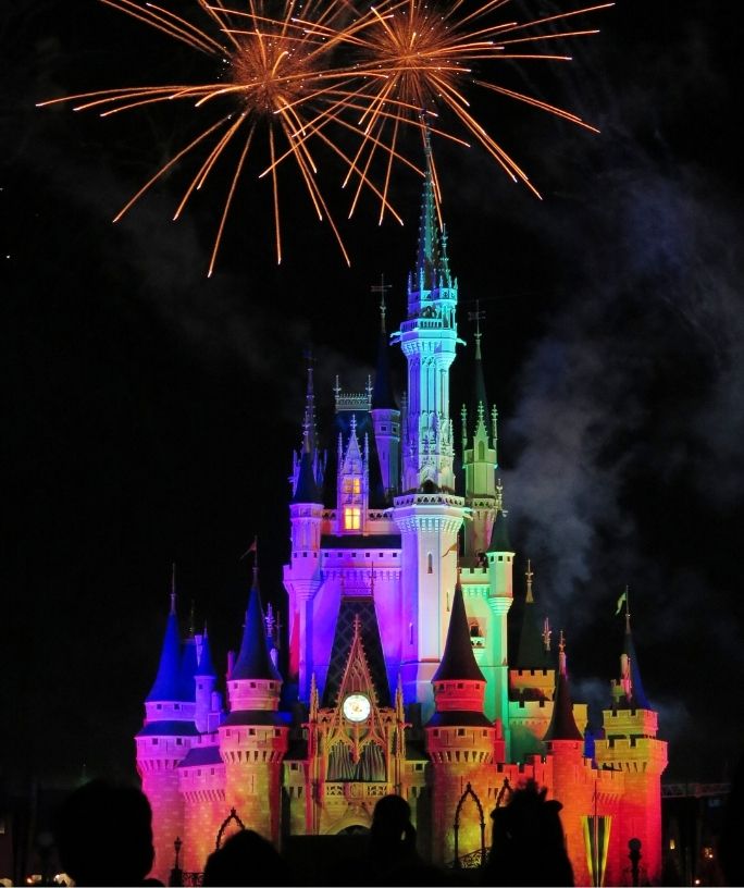 Cinderella's castle in Magic Kingdom at Disney World is lit up with multiple colors as fireworks explode overhead