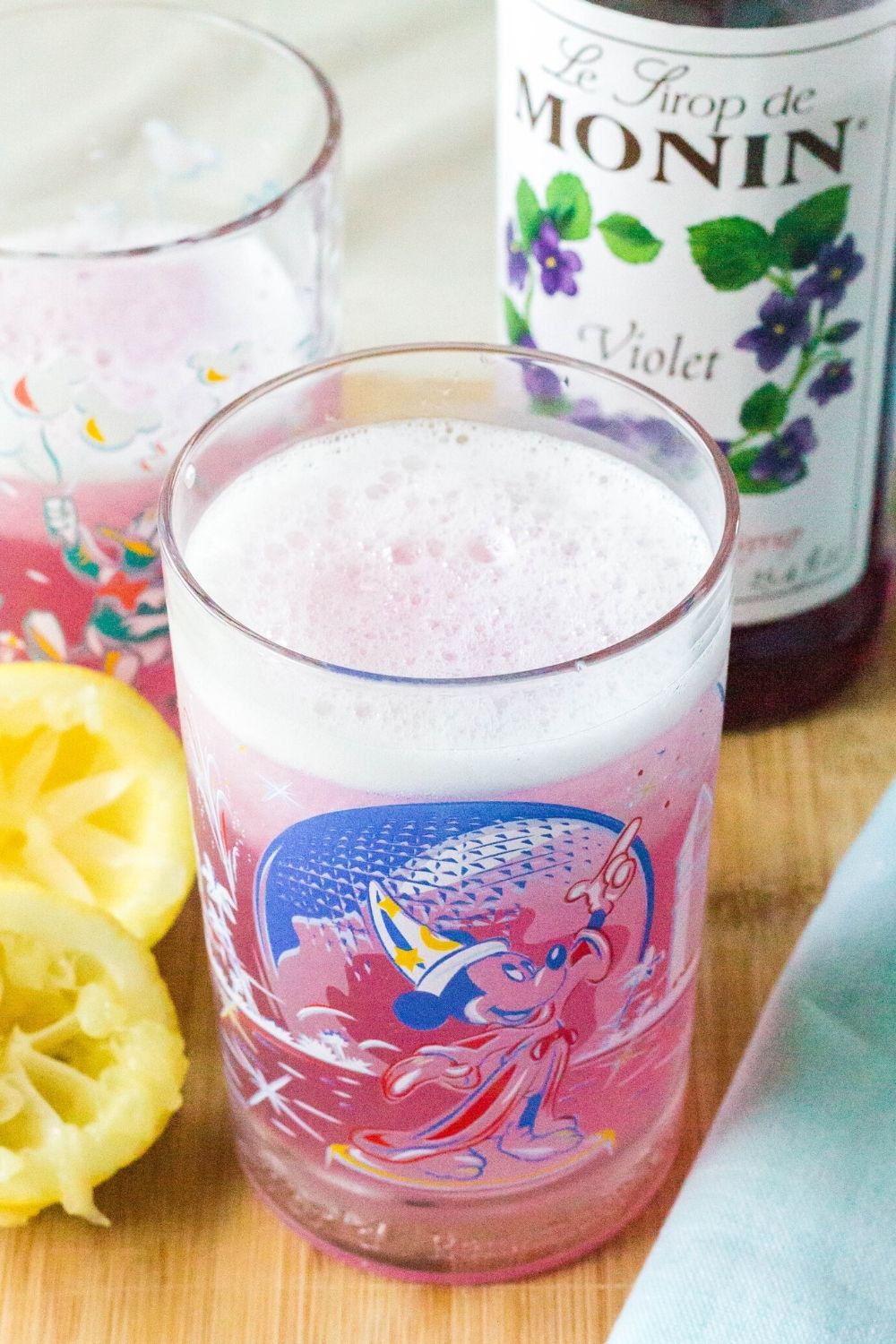 epcot glass filled with purple lemonade, next to squeezed lemons and a bottle of Le Sirop de Monin Violet syrup.