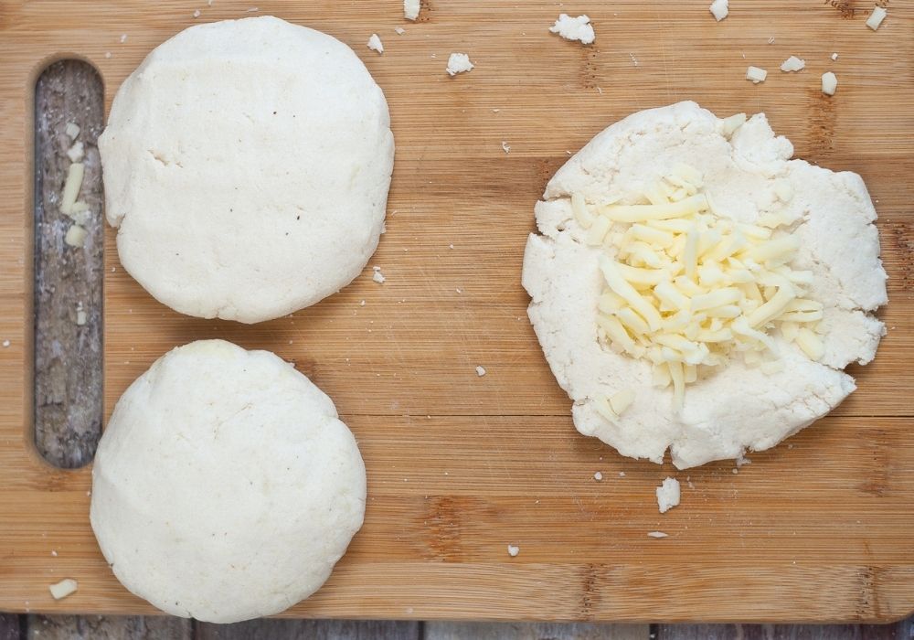rounds of dough prepared into arepas. One arepa is open, with shredded cheese in the center, waiting to be closed into a patty for cooking.
