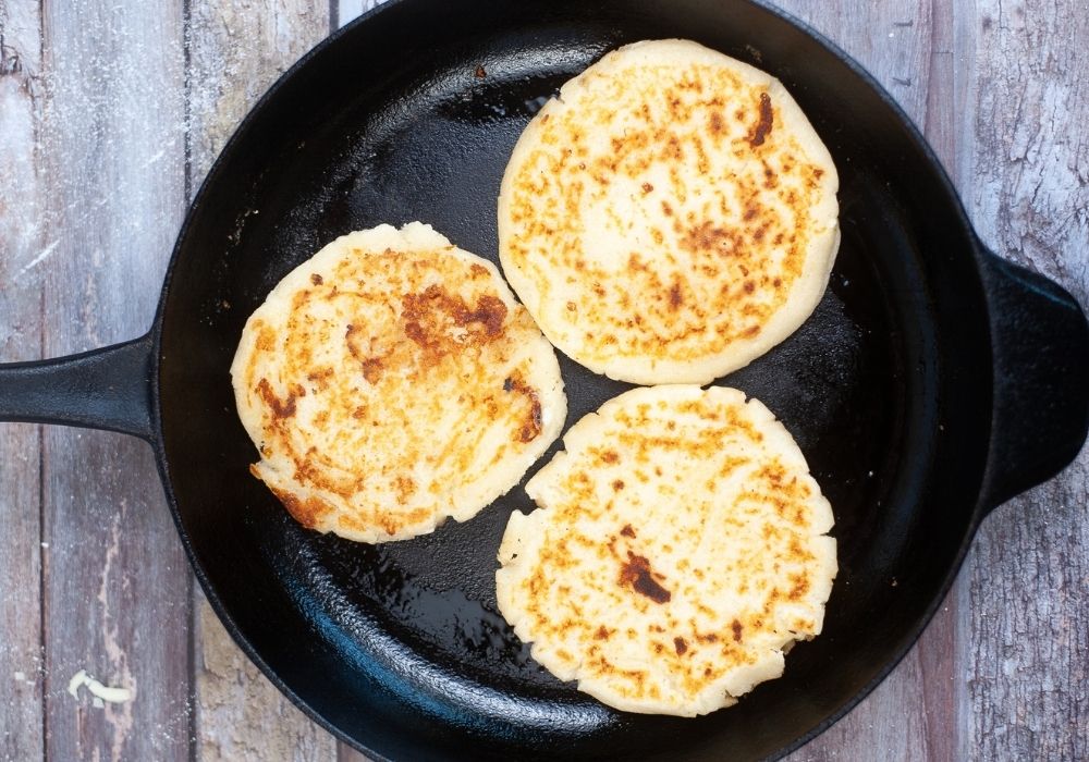 three arepas con queso being cooked in cast iron skillet