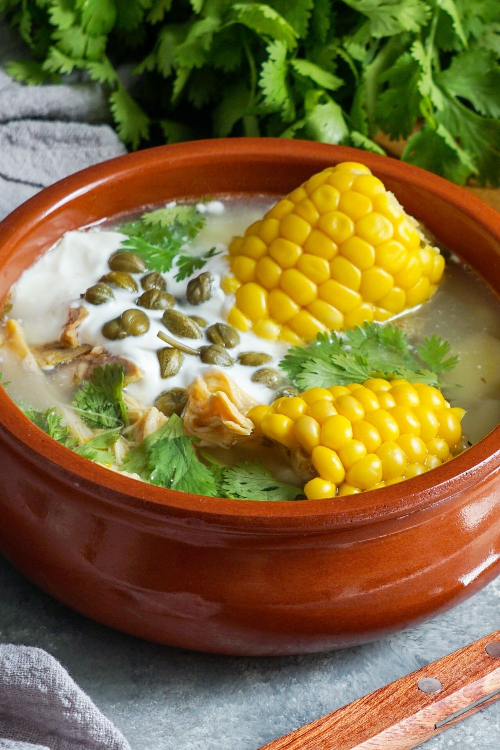 close-up view of an orange bowl filled with ajiaco colombiano, including chicken, corn on the cob segments, potatoes, cilantro, cream, and capers